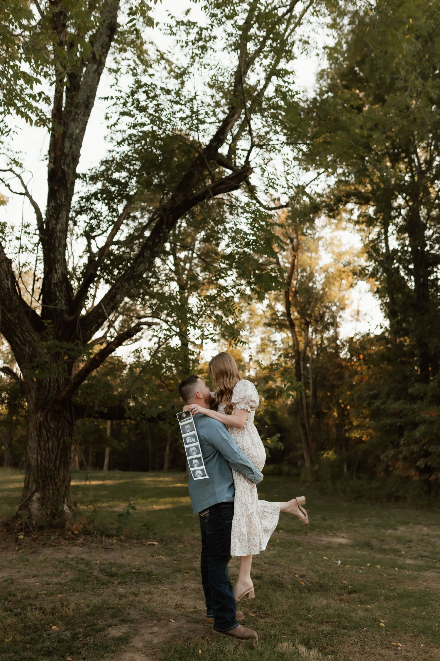Man lifting woman in white dress outdoors during golden hour in lush green park setting.