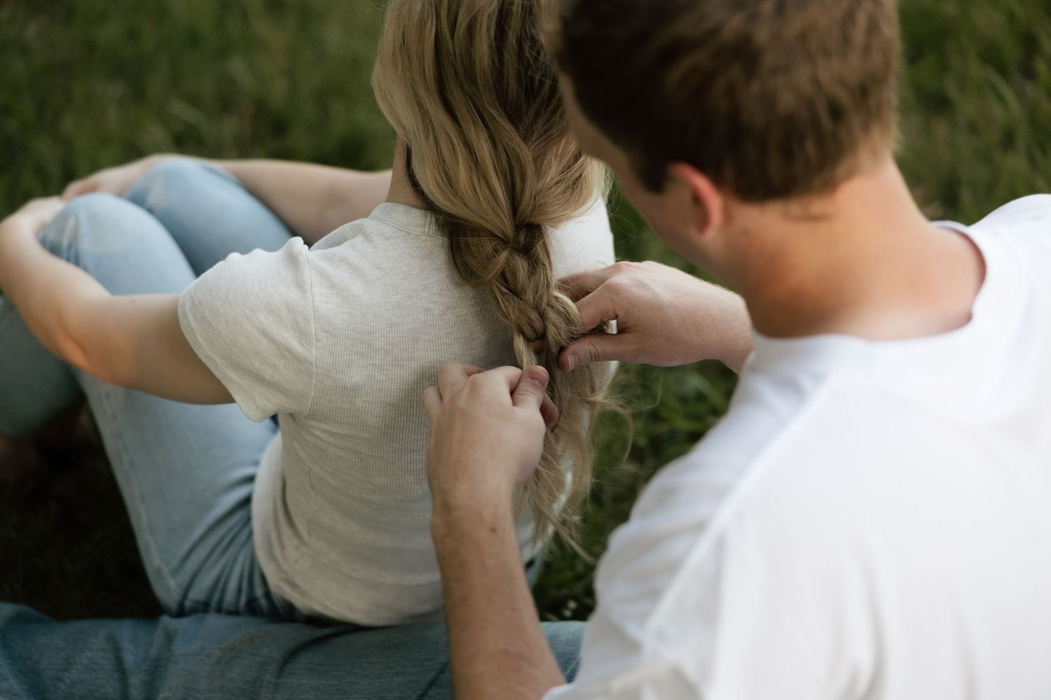 A man braids a blonde woman's hair as they sit together on grass during a romantic outdoor photo session.