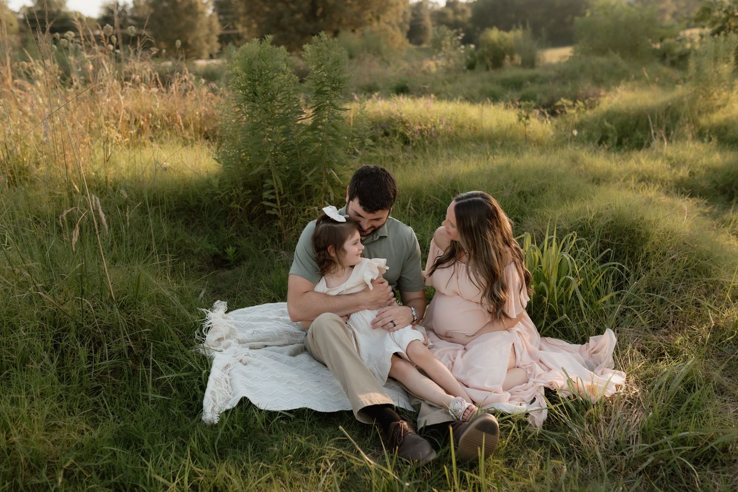 Pregnant woman, man, and young daughter sitting together on blanket in tall green grass at sunset.