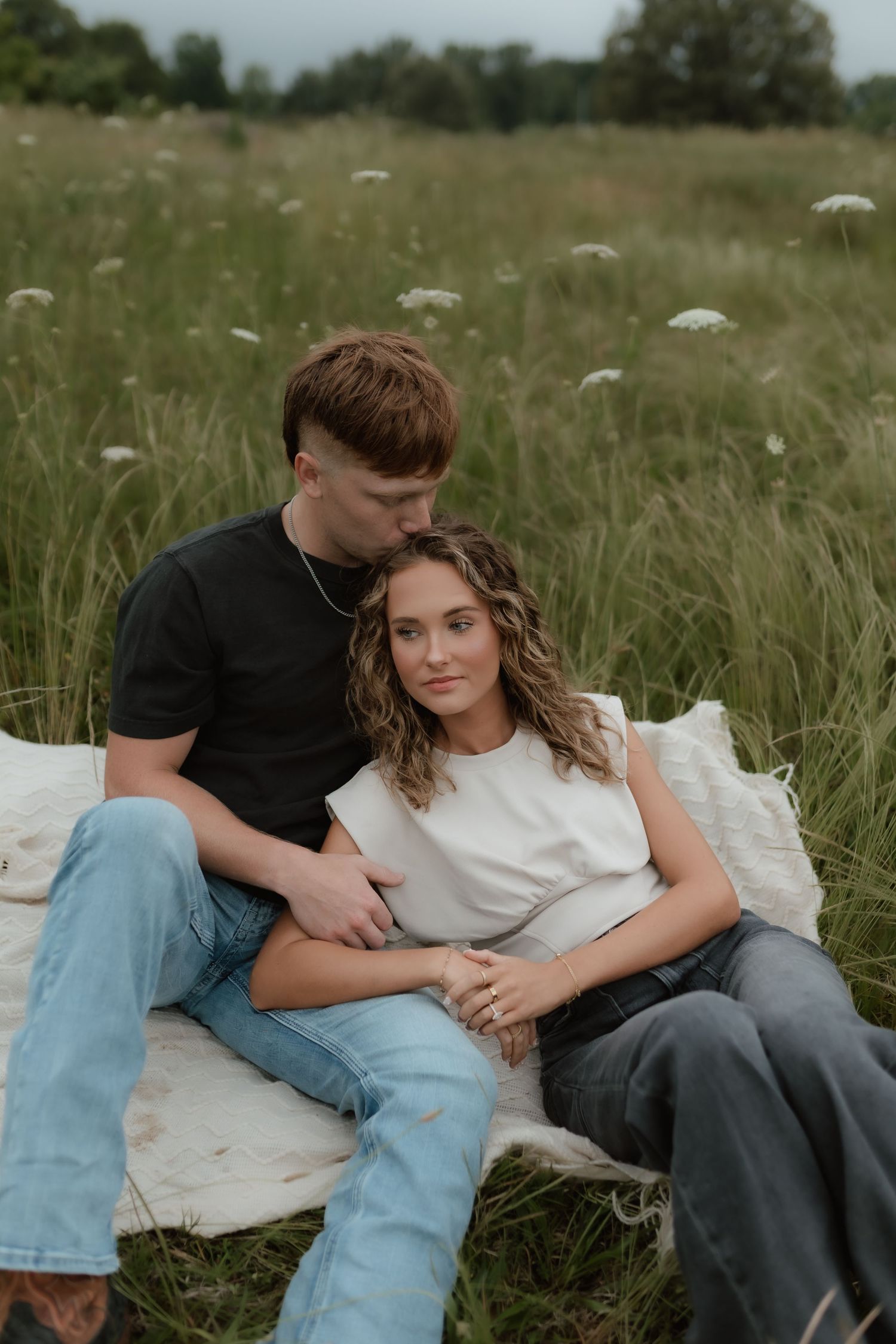 Couple sitting on white blanket in green field, man kissing woman's head while she leans against him.