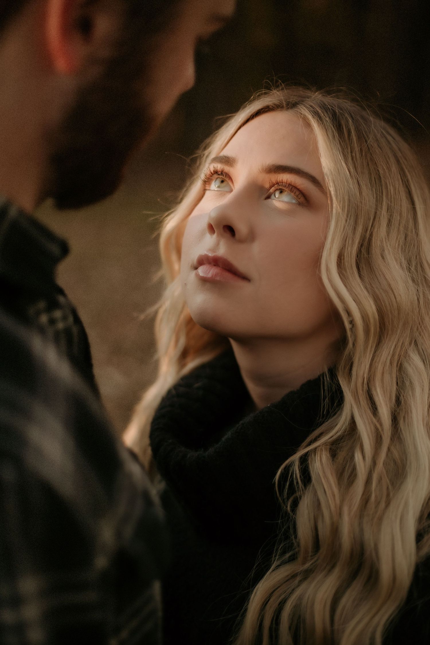 Blonde woman gazes up lovingly at bearded man in warm golden light during romantic outdoor couples session.