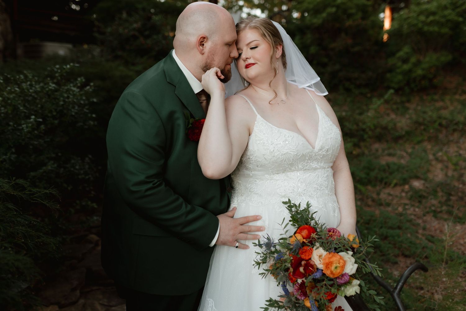 Groom in green suit tenderly kisses bride holding colorful bouquet in lush garden setting.