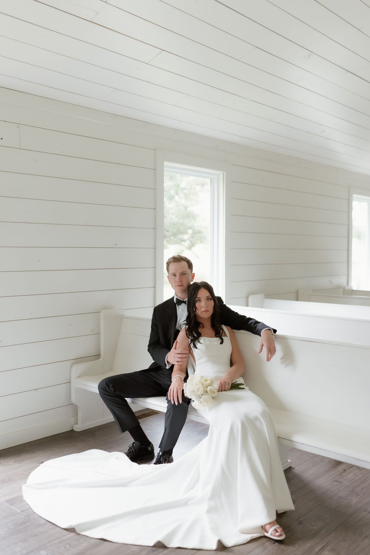 Elegant bride and groom posing together inside a white shiplap chapel with bouquet of white flowers.