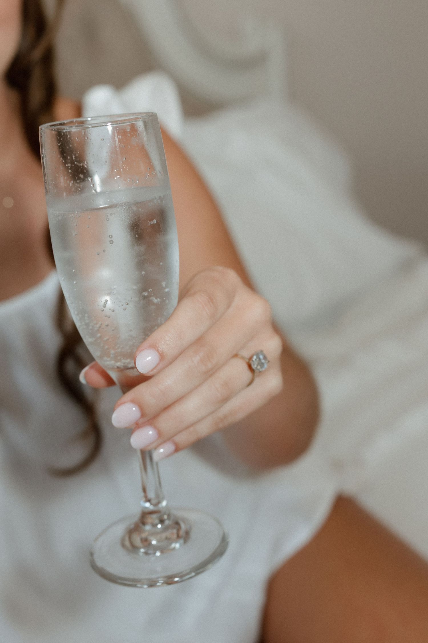 Bride wearing engagement ring holds a champagne flute in a white robe before wedding.