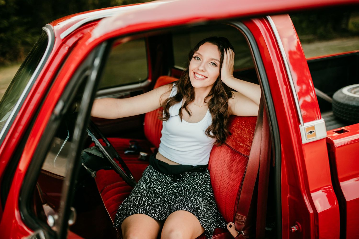 Smiling young woman in white crop top sits inside red vintage truck with hands framing her face.