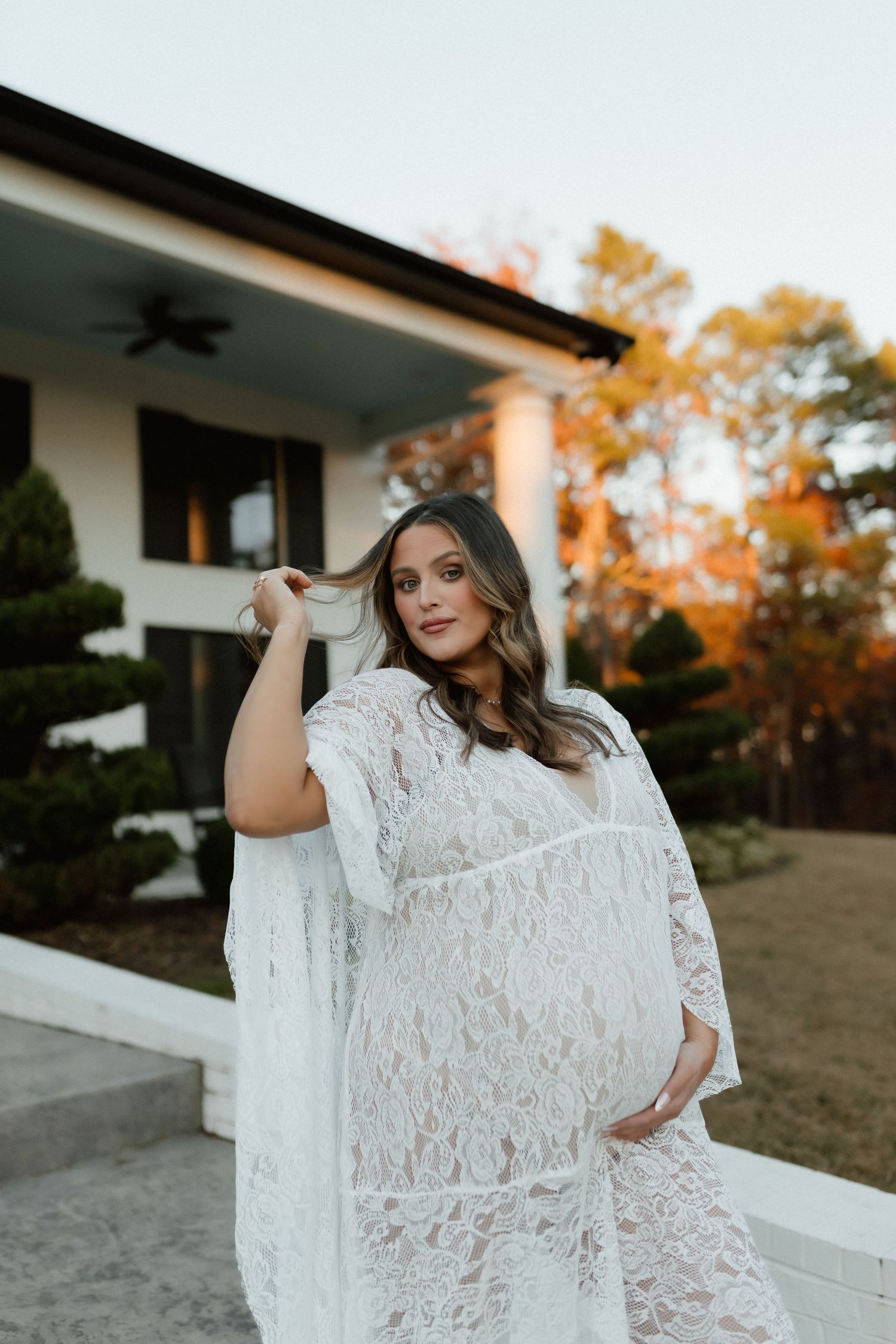 Pregnant woman in white lace maternity gown posing outdoors at sunset in front of modern white home.