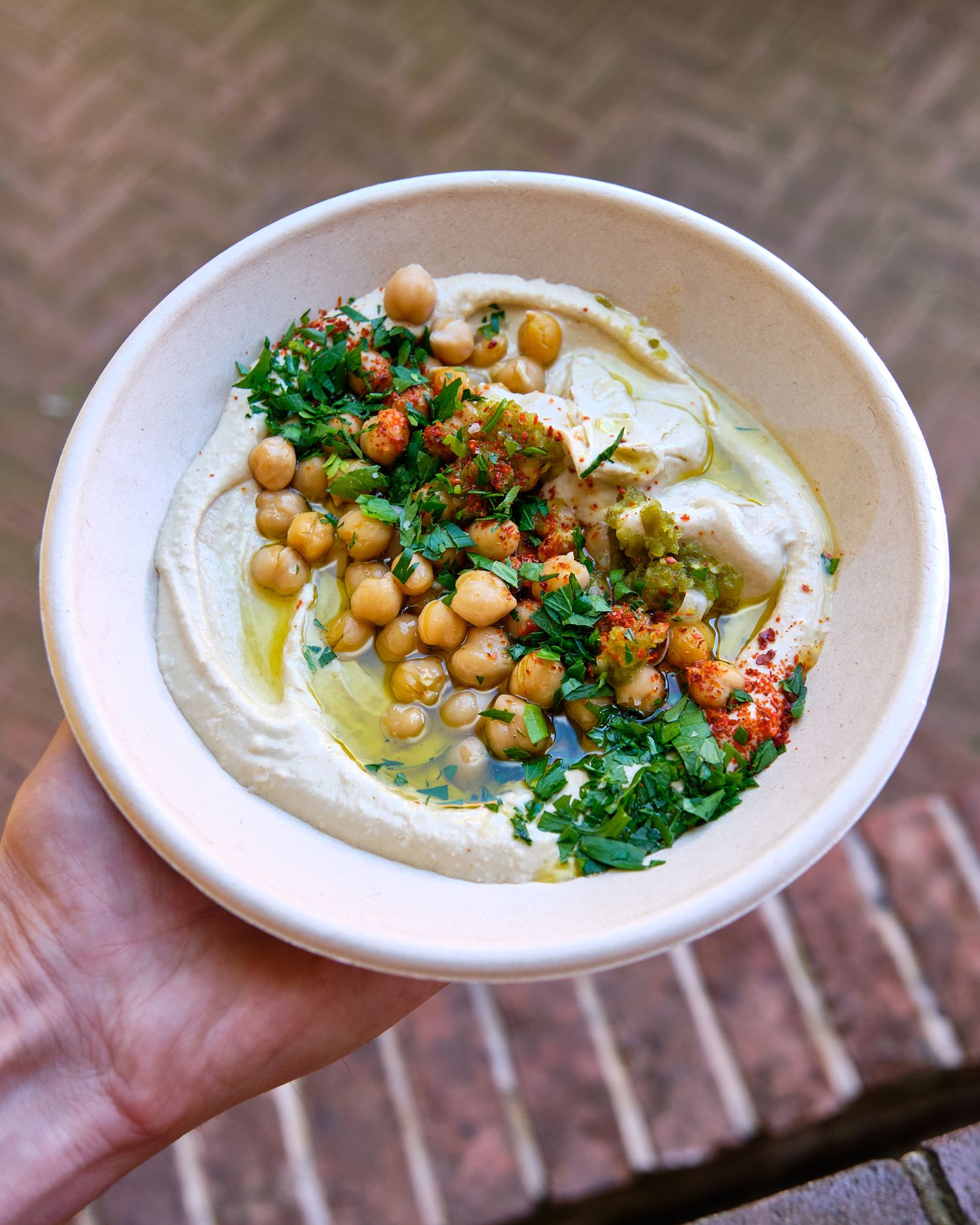 Bowl of smooth hummus garnished with chickpeas, parsley and spices on brick background.
