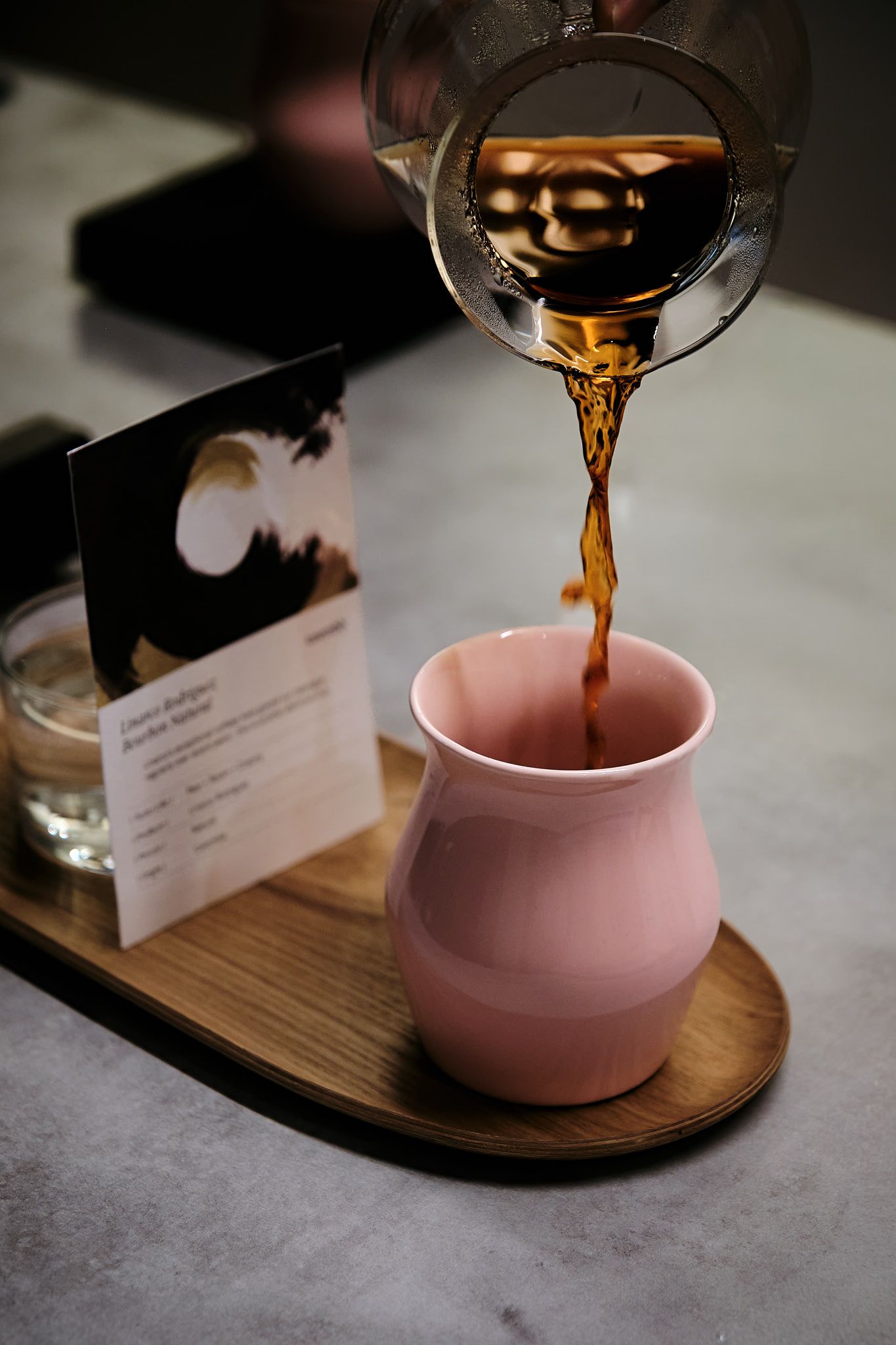 Coffee cascades from glass carafe into pink ceramic cup with menu card visible.