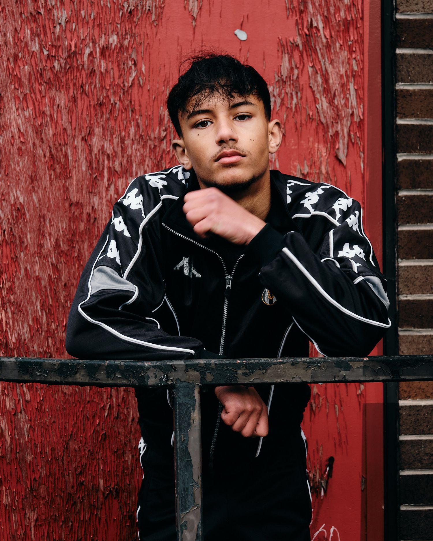 Fashion portrait showcasing black Kappa sports jacket against vibrant red wall.