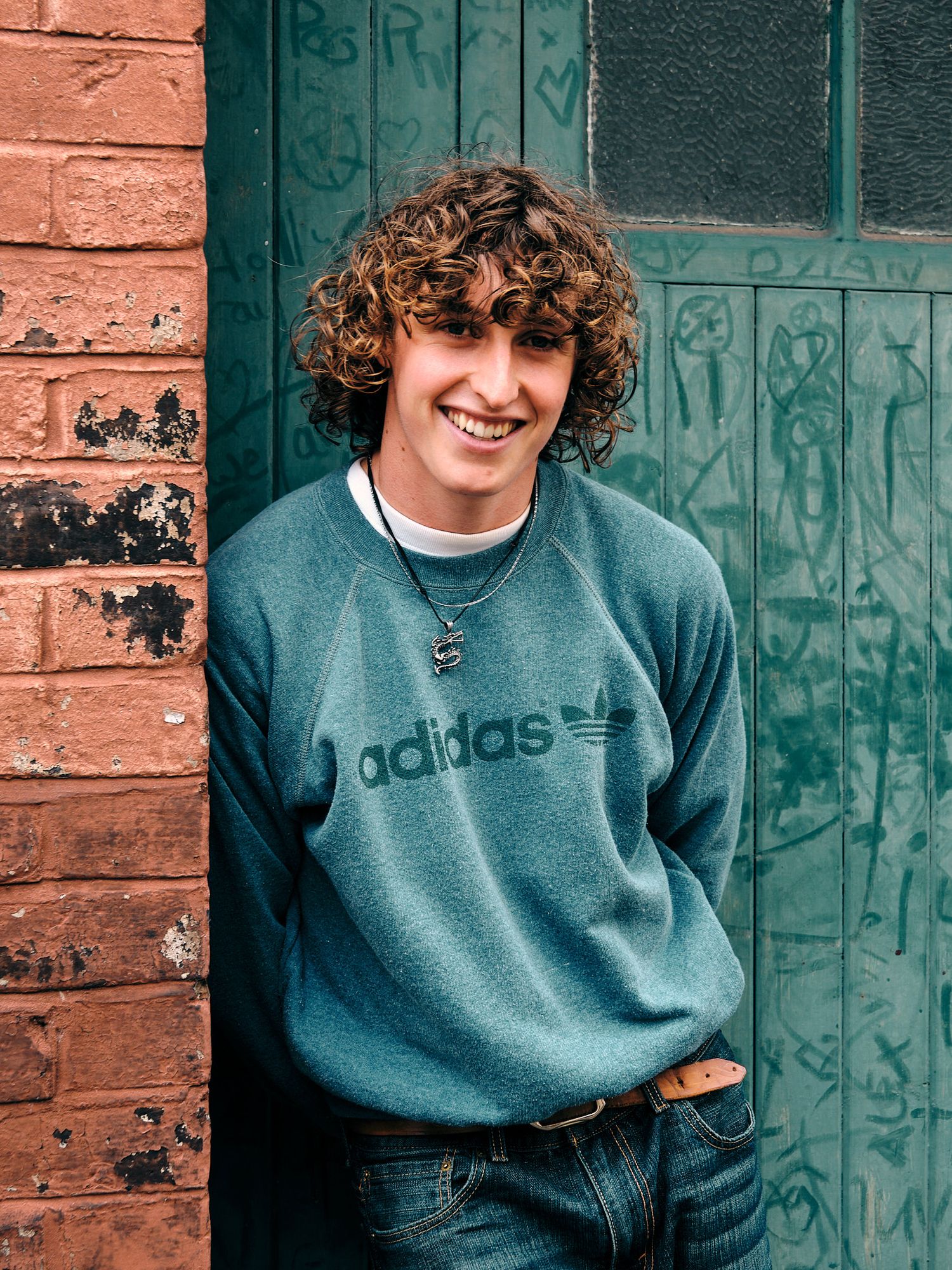 Person wearing blue Adidas sweatshirt sitting against brick wall with green door.