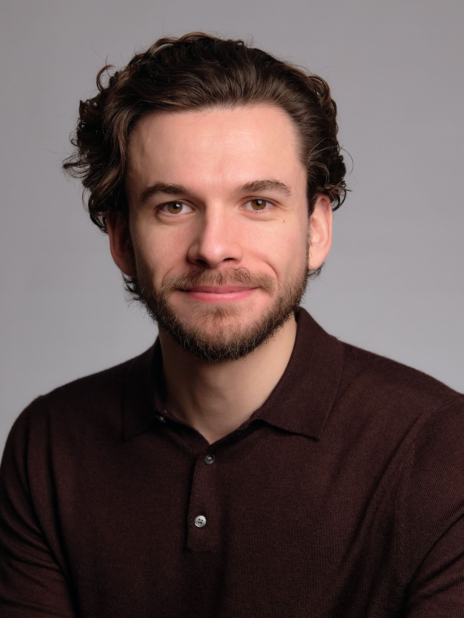 Professional headshot portrait of someone wearing a brown sweater against a neutral gray background.