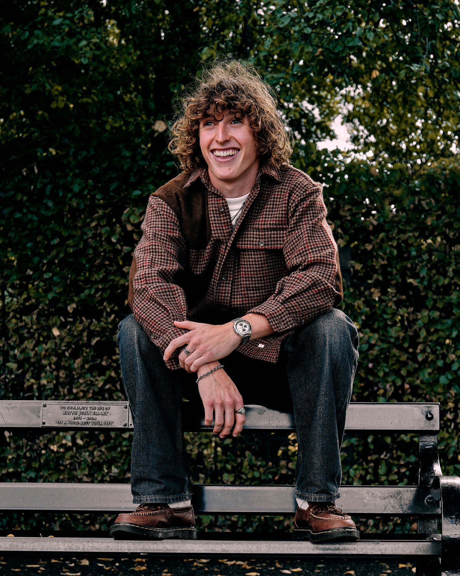 Person in brown plaid jacket sitting relaxed on park bench with greenery background.
