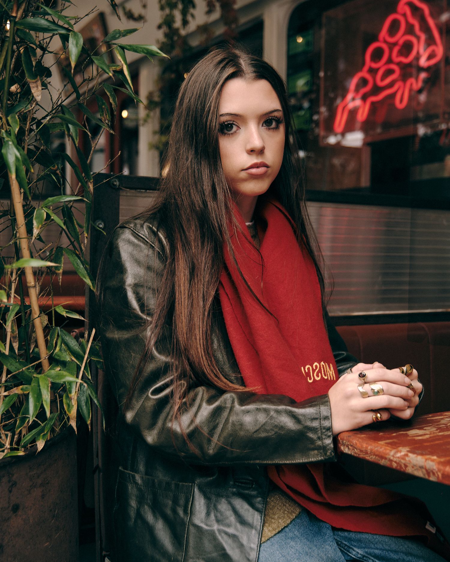 Person in red scarf and leather jacket sitting at cafe with neon signs visible.