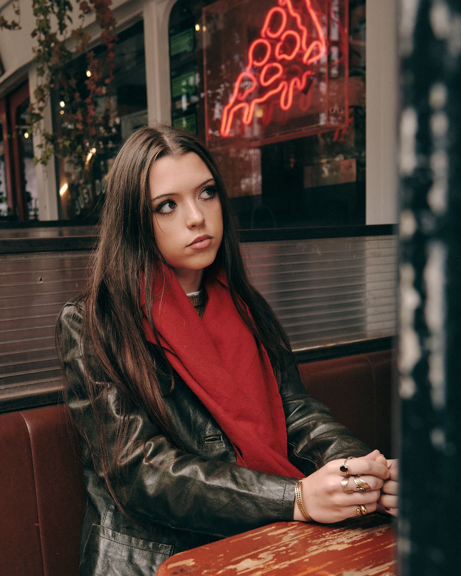 Person in red scarf and leather jacket sitting at cafe table with neon signs.