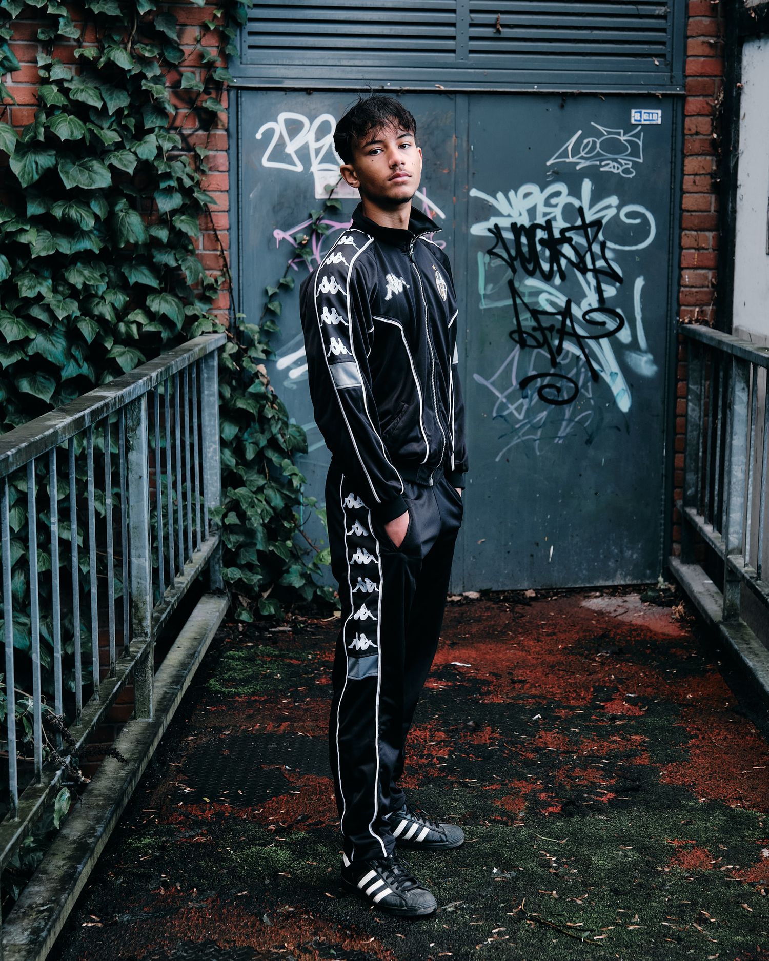 Model in black tracksuit poses against graffiti wall and ivy backdrop in urban setting.