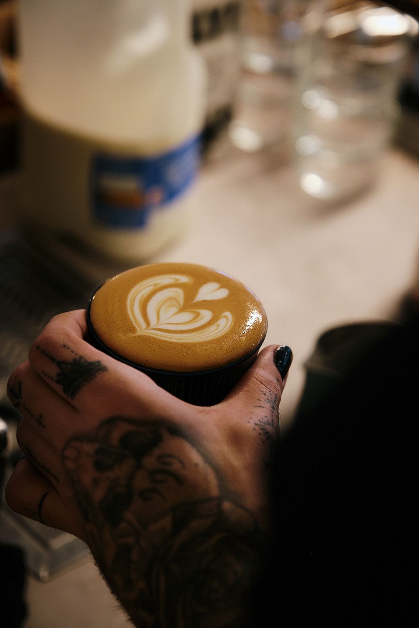 Detailed latte art heart pattern in coffee cup held by tattooed hands.