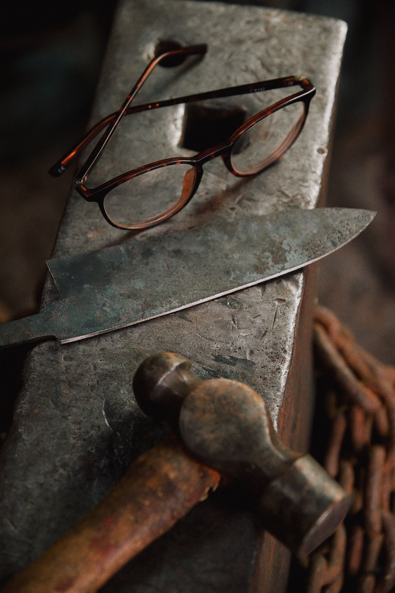 Vintage tortoiseshell glasses rest on a concrete surface beside an old hammer and knife in moody lighting.