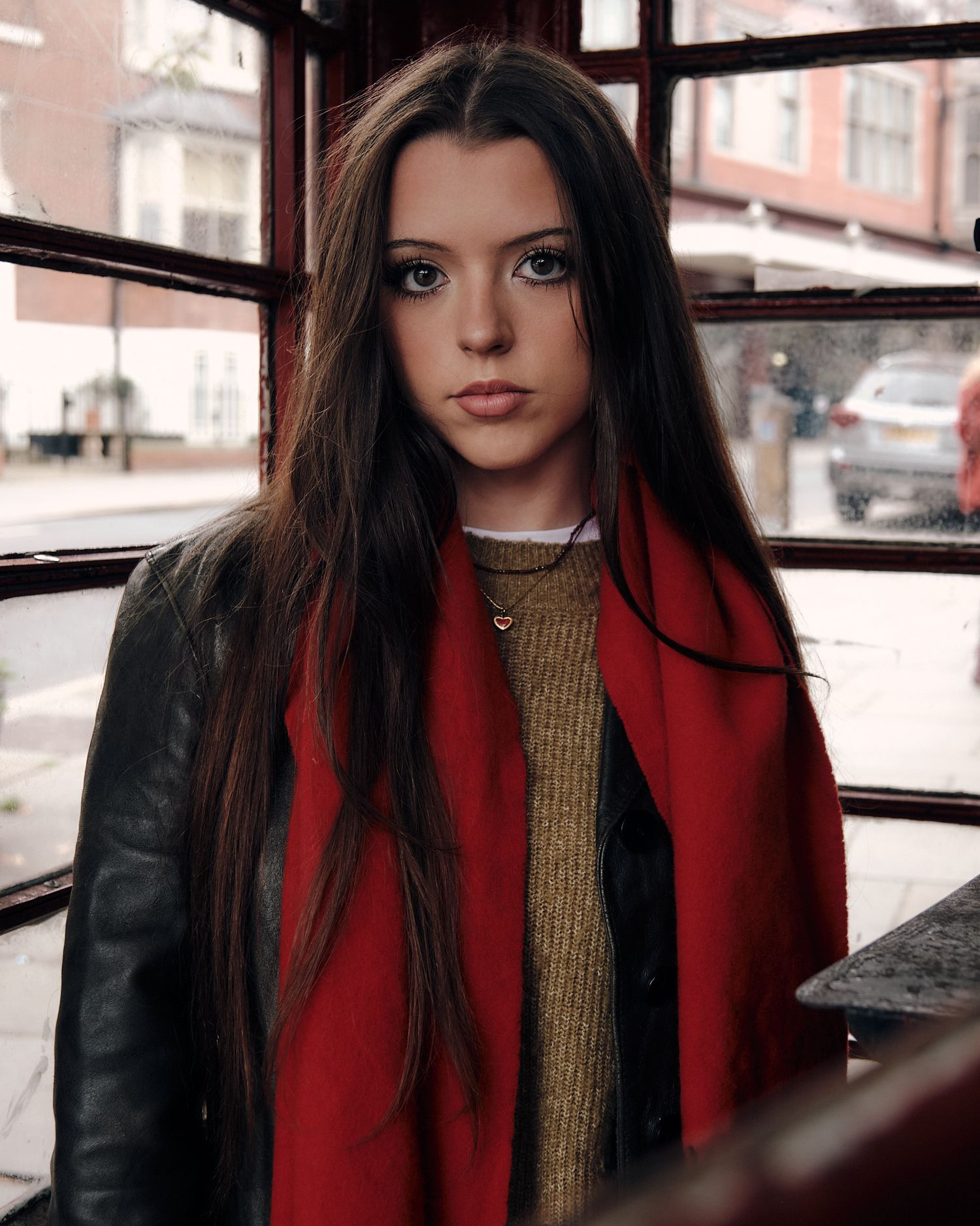 Person wearing red scarf and dark jacket sitting by window in urban setting.