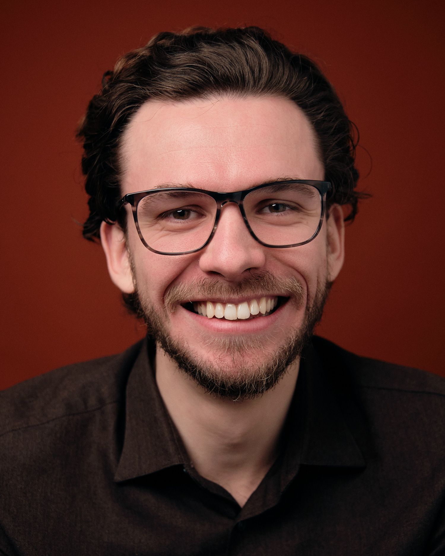 A series of headshots against a dark red background showing someone wearing glasses with curly hair and facial hair smiling.