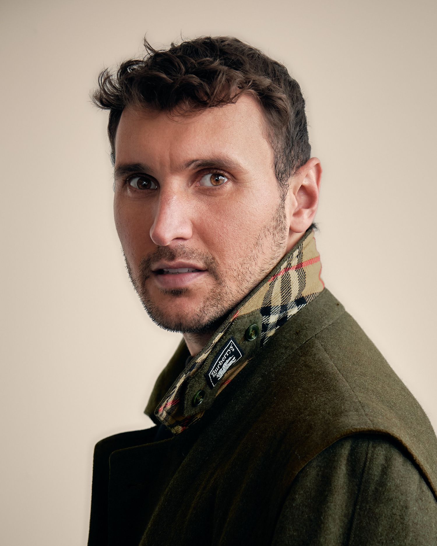 A person wearing a dark olive green coat poses against a beige background in a series of profile and angled shots.