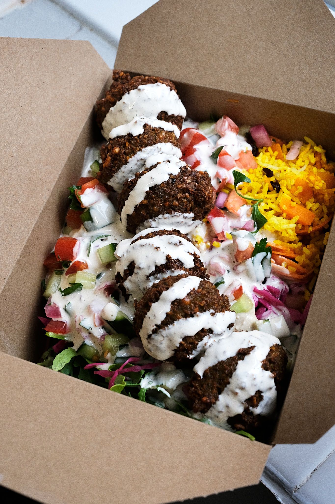 Middle Eastern falafel bowl with creamy sauce and colorful toppings arranged in eco-friendly takeout box.