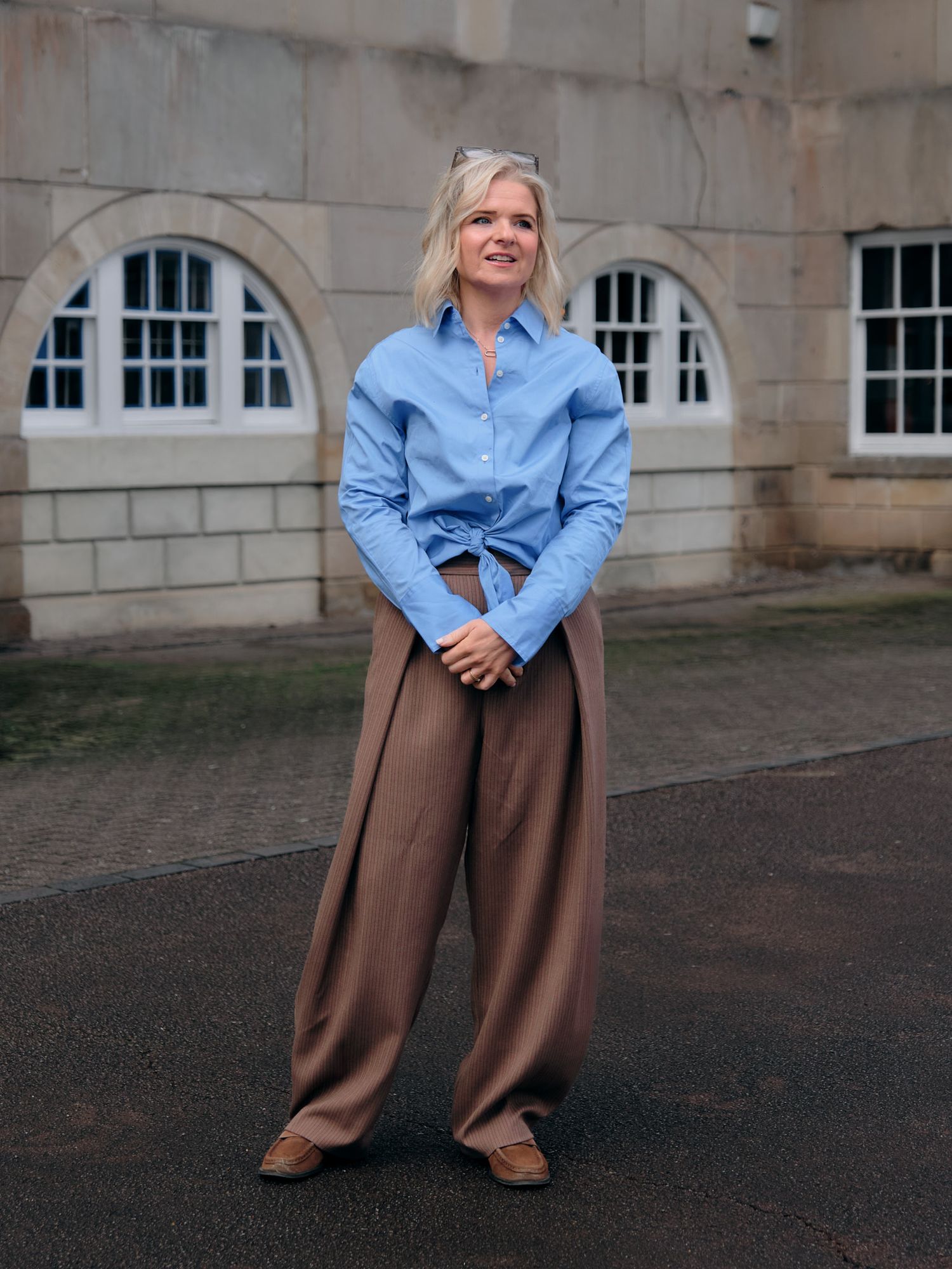 A fashion model poses in brown wide-leg trousers and a cropped blue button-down shirt against a stone building exterior.