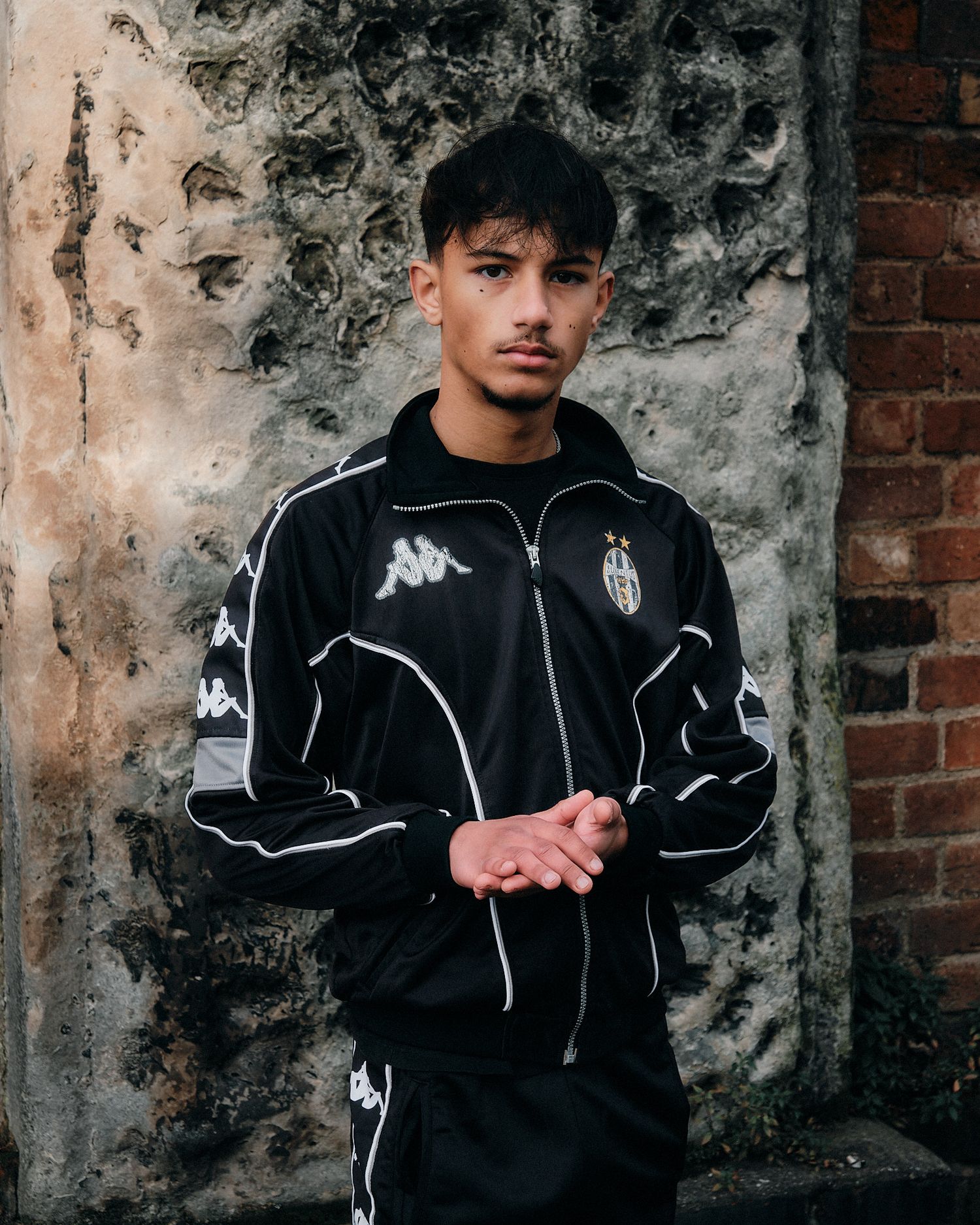 Casual portrait featuring black Kappa sports jacket against weathered brick wall.