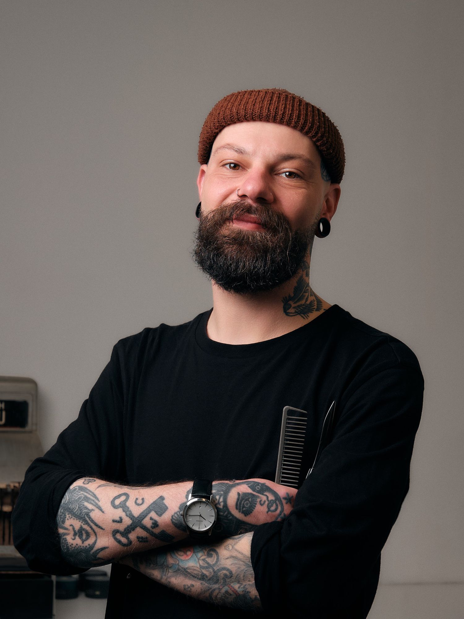 Bearded hairstylist in brown beanie and black sweater with visible tattoos stands confidently in salon setting.