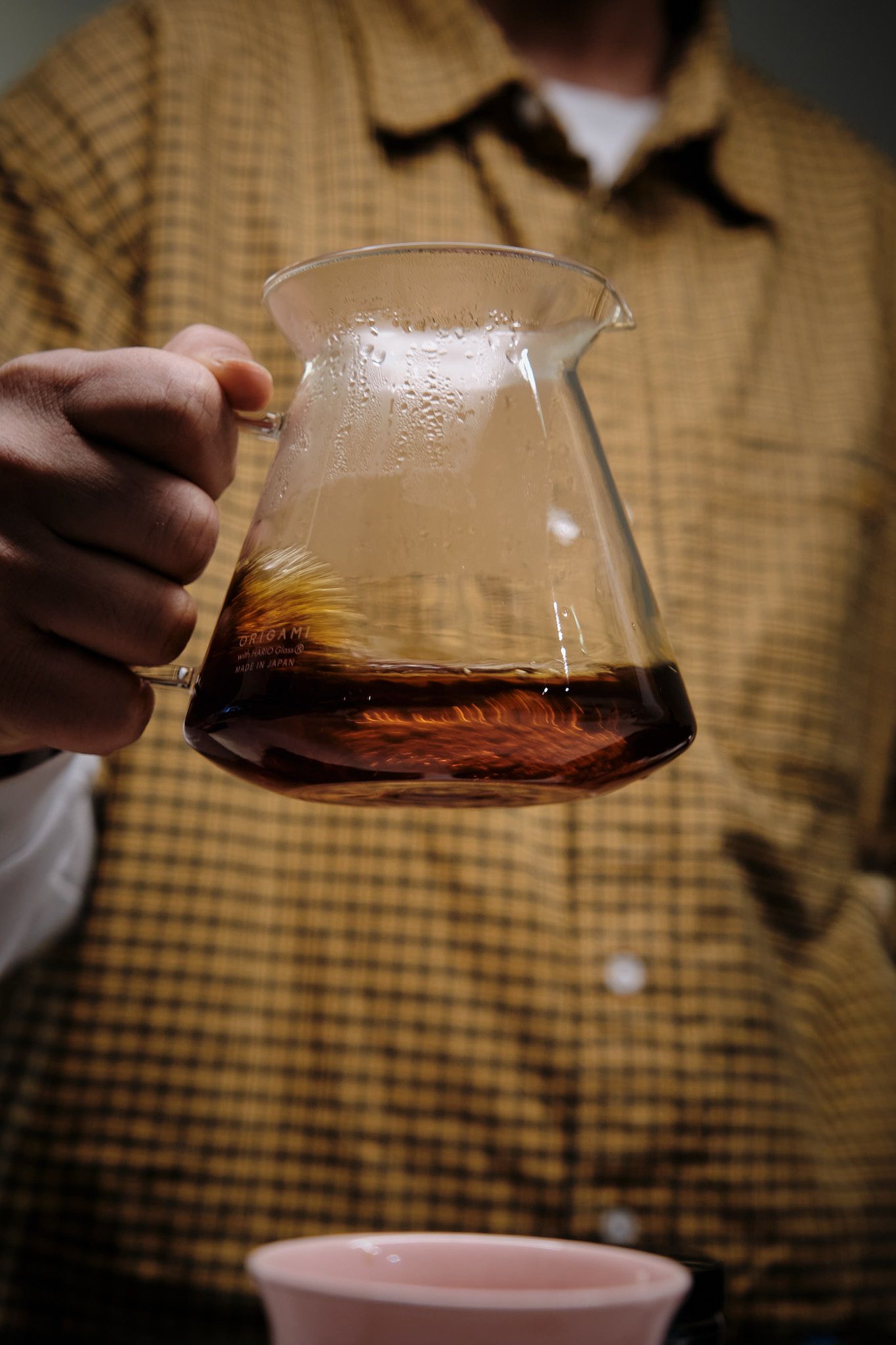 Glass coffee carafe held against checkered shirt showing amber liquid inside.