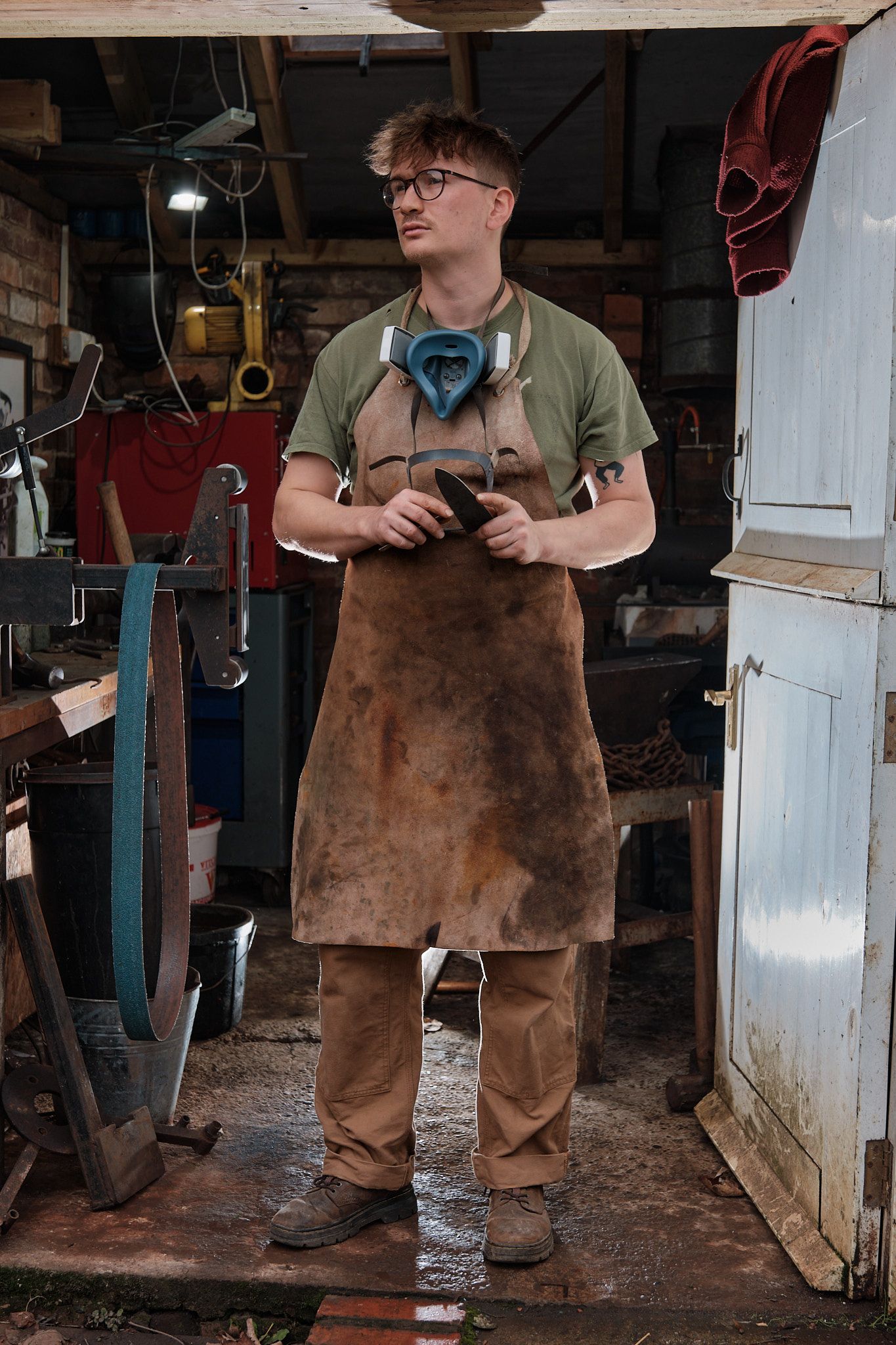 Craftsperson wearing brown leather apron and safety glasses works in rustic workshop environment.