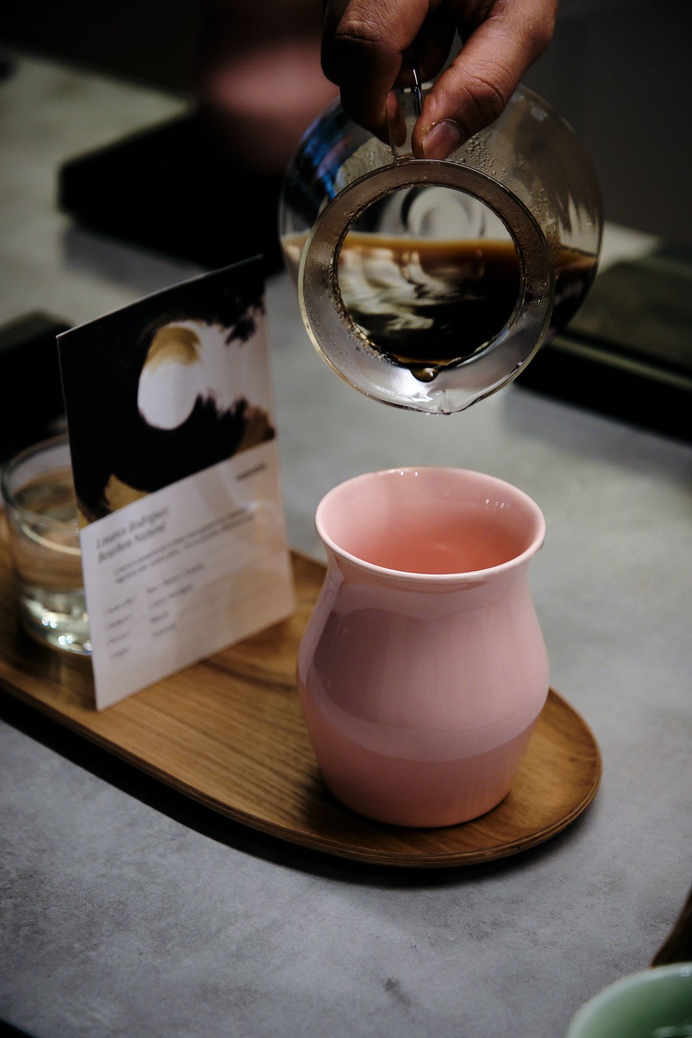 Hot coffee being poured into a pink ceramic vessel on a wooden coaster.