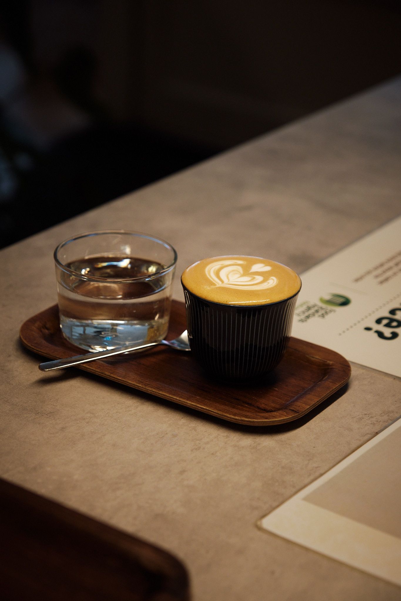 Latte with heart design and glass of water served on wooden serving board.