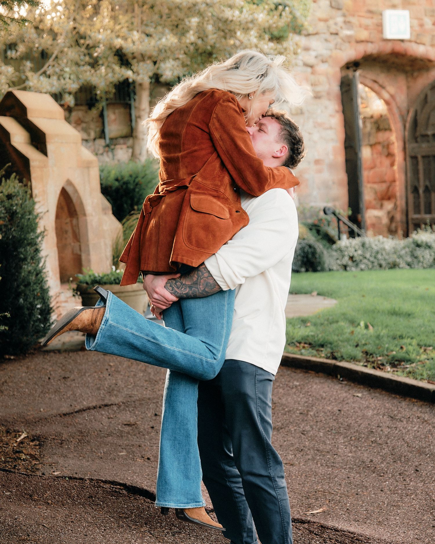 Playful embrace between couple near brick architecture and garden path.