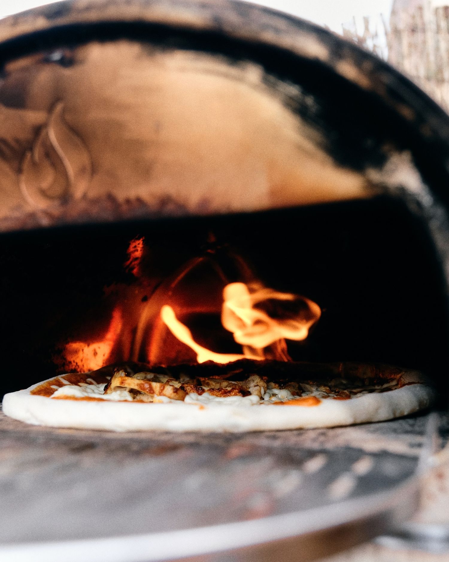 Wood-fired pizza cooking in a traditional brick oven with flames visible.