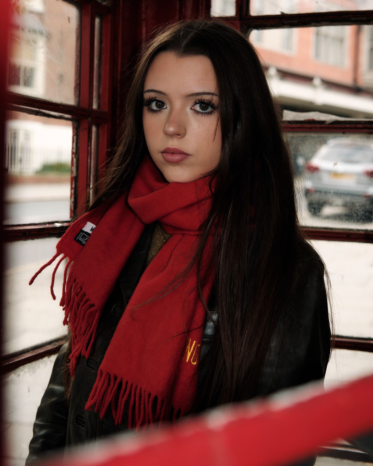 Dramatic portrait inside red phone booth showcasing bright red scarf and dark winter fashion.