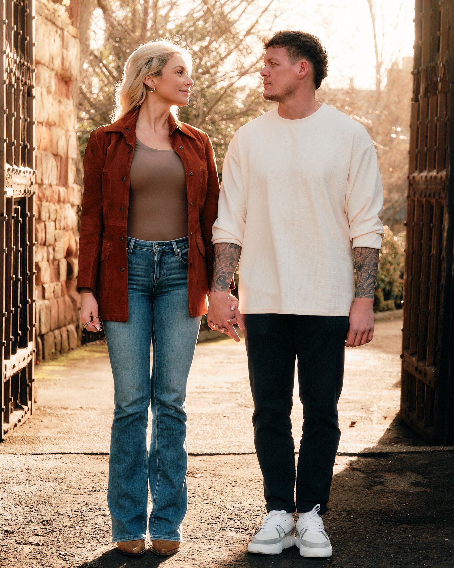 Couple walking hand in hand through stone archway during golden hour in casual outfits.