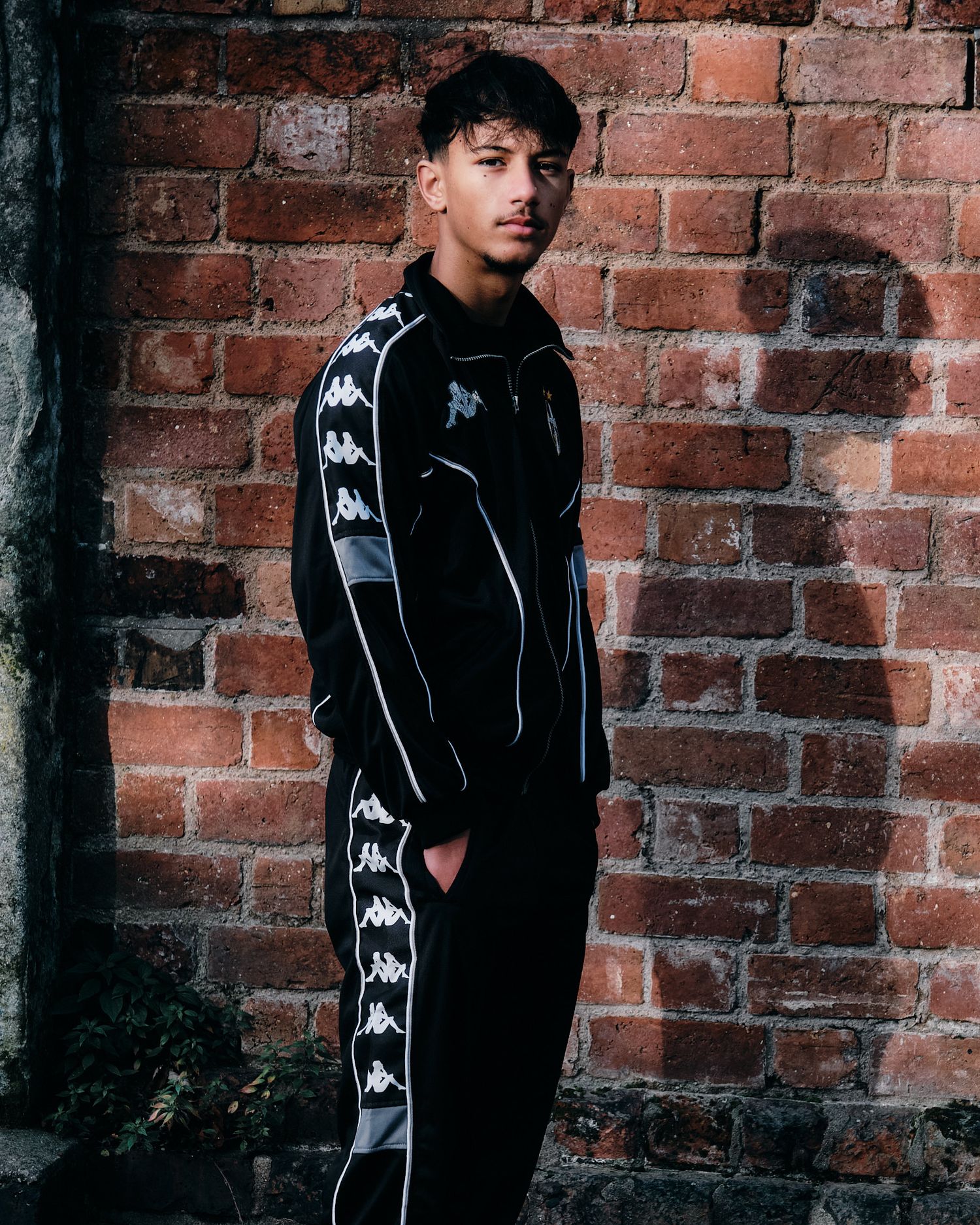 Athletic wear photo showing full black Kappa tracksuit against brick wall background.