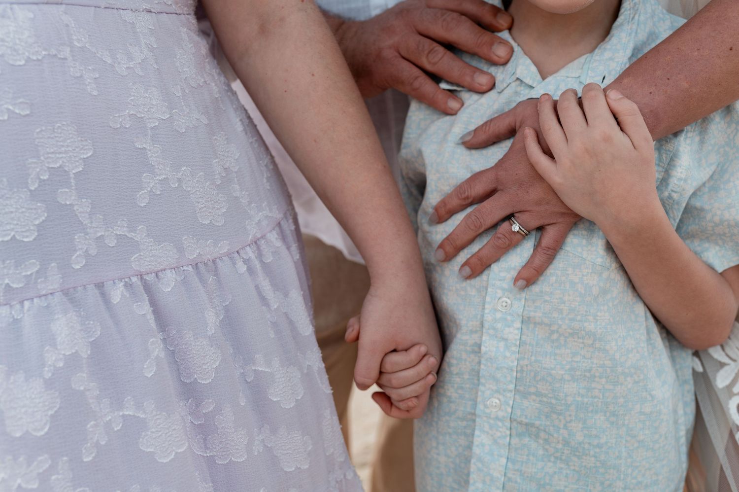 Adult hands lovingly embrace a child in a light blue outfit while holding hands with a woman in a white lace dress.