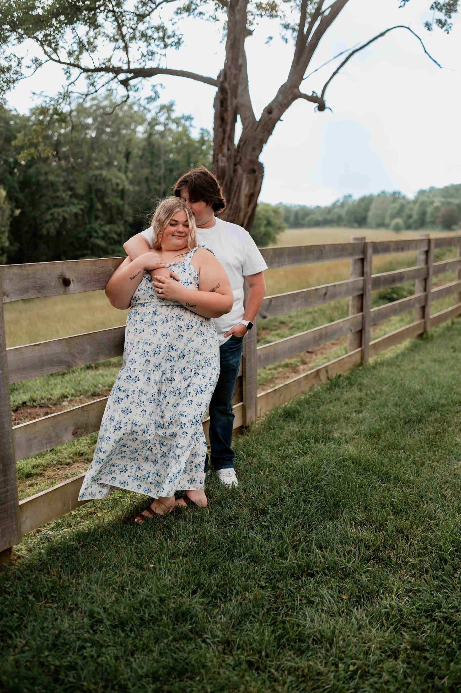 Couple sharing a romantic moment on grassy hillside with wooden fence in background.