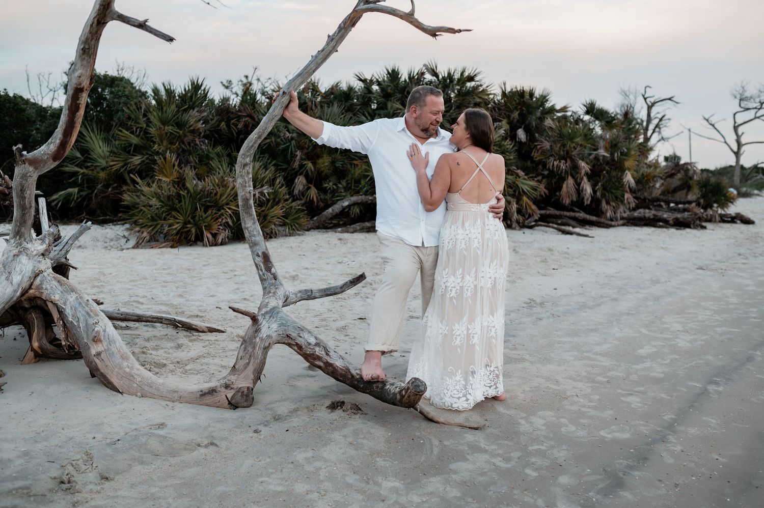 Person in flowing white dress posing on sandy beach with driftwood.