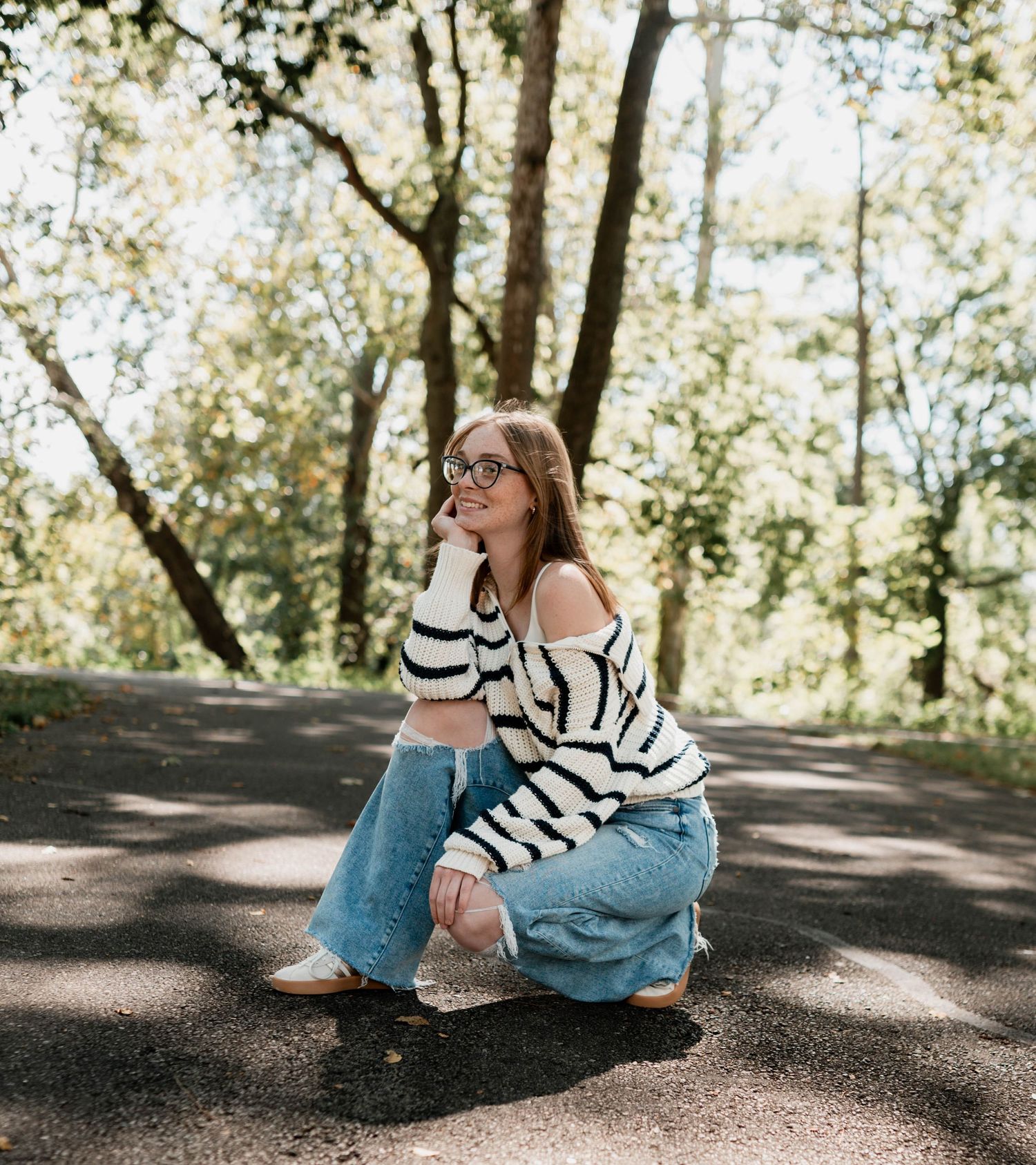 Someone in denim and white sweater sitting on ground in dappled sunlight.