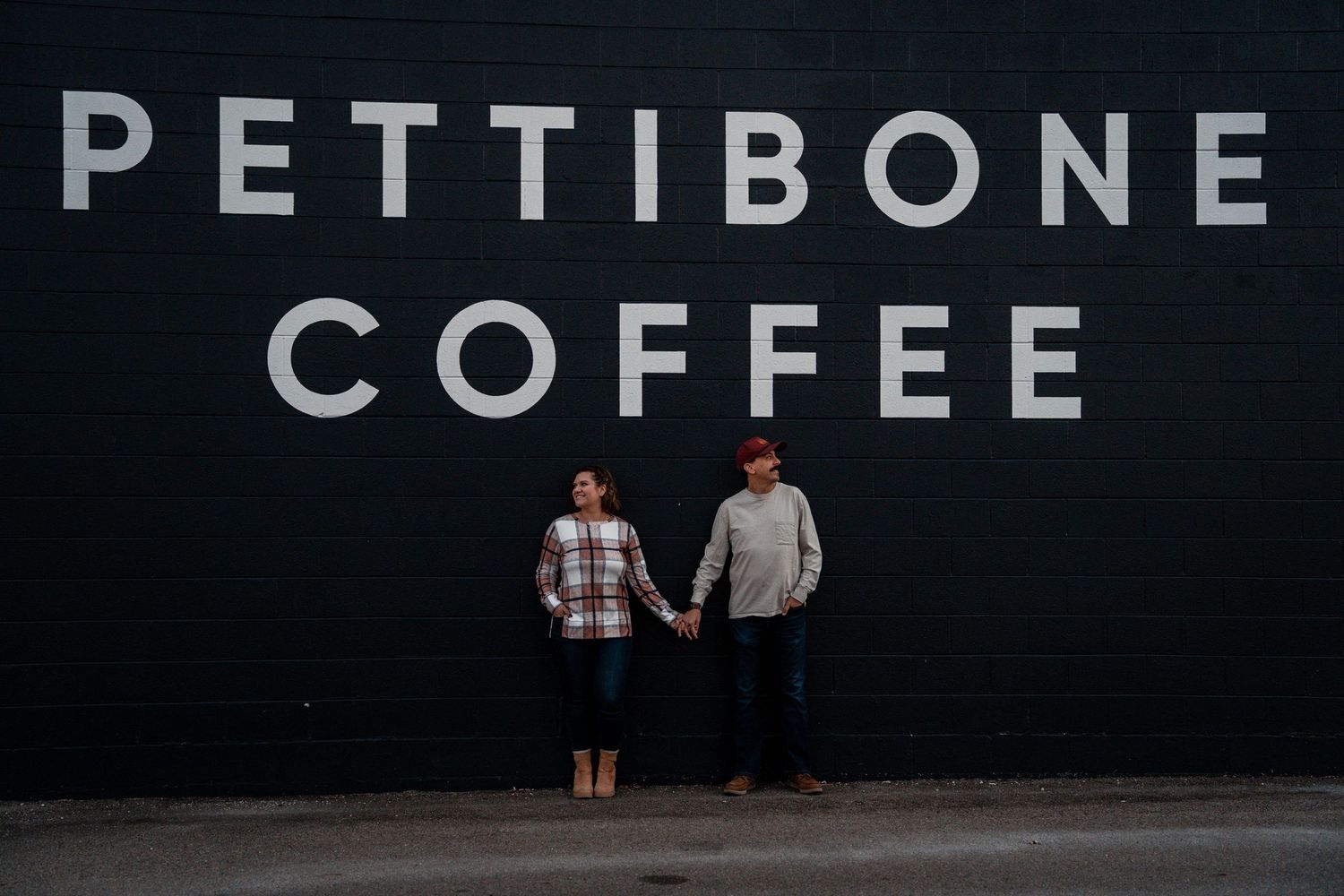 Couple holding hands in front of Pettibone Coffee black wall sign, casual engagement photo session.