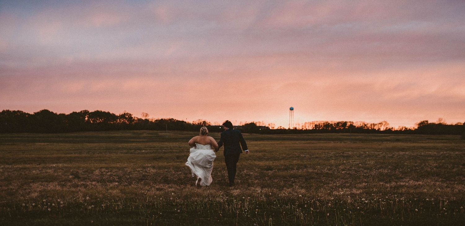 Bride and groom running hand in hand across a field at sunset with a vibrant pink and purple sky backdrop.