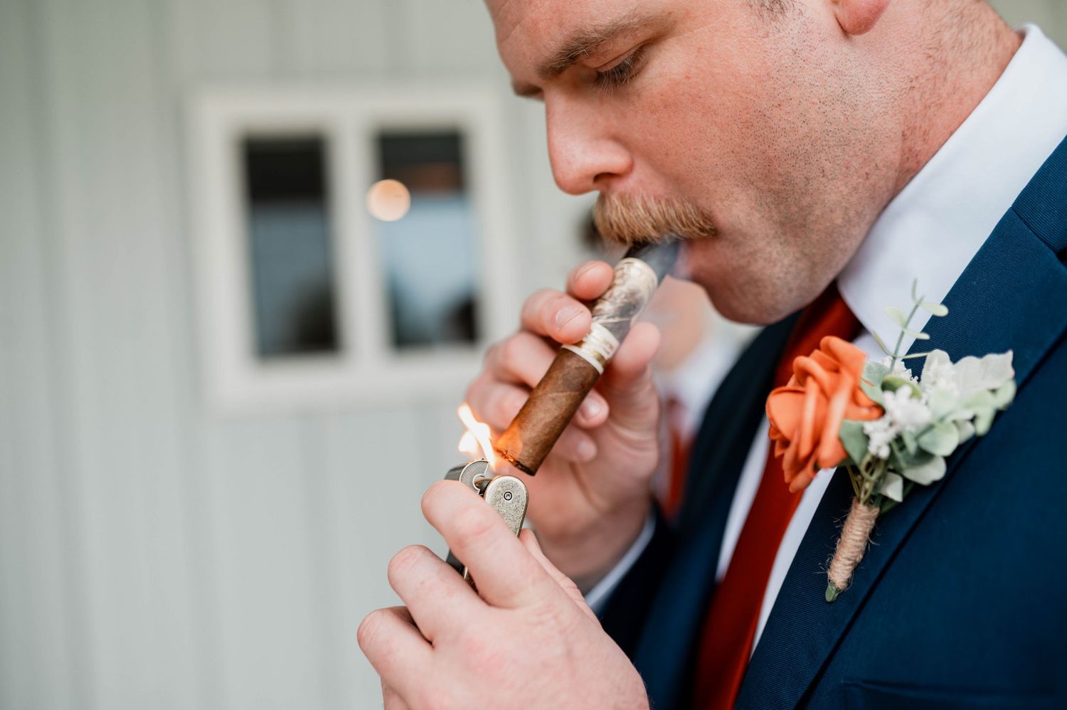 Groom in navy suit lighting a cigar with orange rose boutonniere on wedding day.