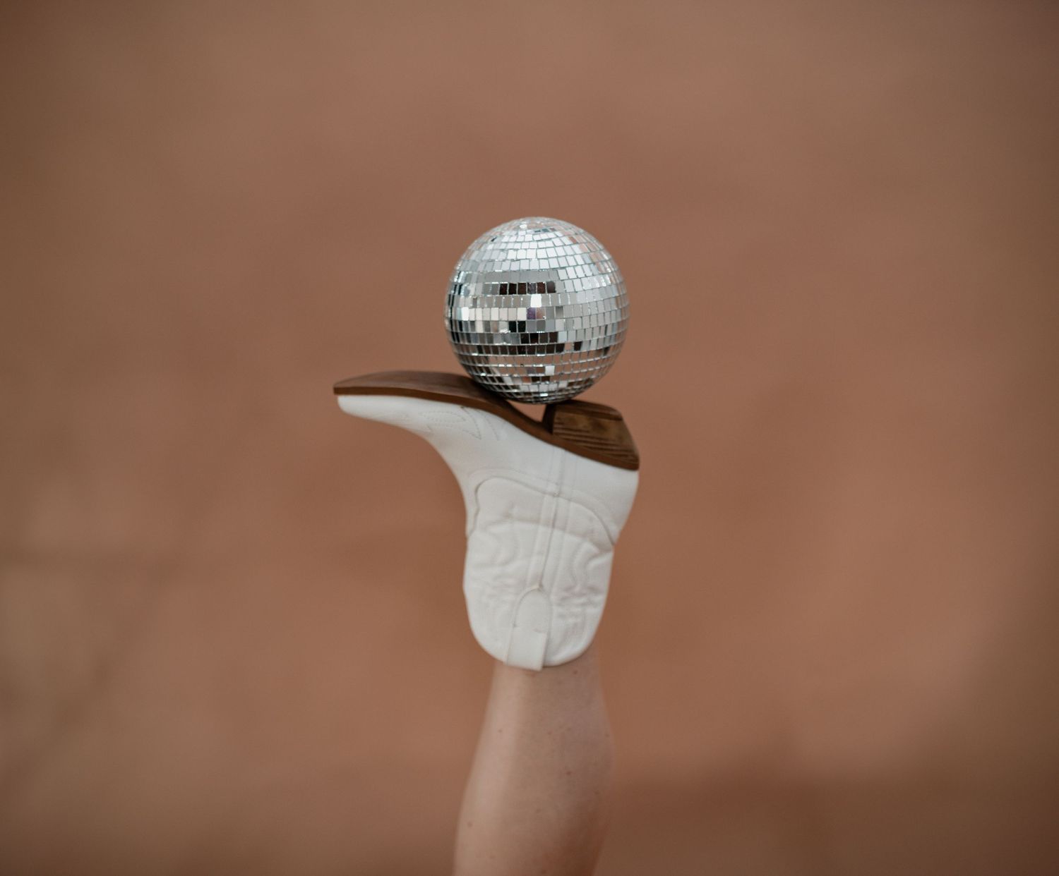 A miniature disco ball balanced on top of a white cowboy boot finger puppet against a beige background.