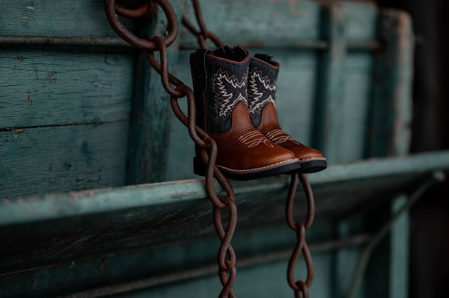 Small brown cowboy boots with black embroidery hanging on a rustic chain against a weathered teal fence.
