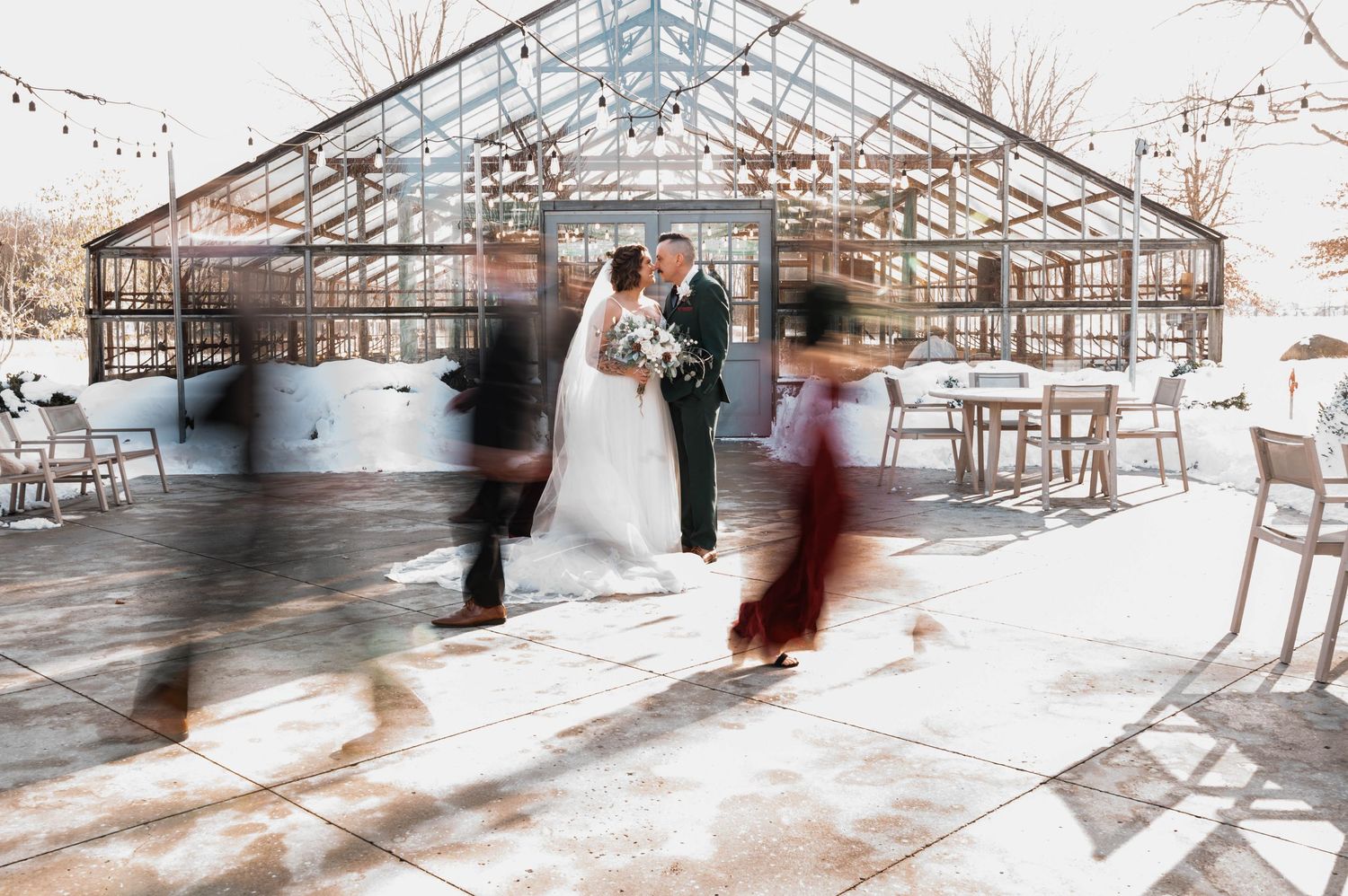 Bride and groom share a kiss outside a glass greenhouse venue while guests move around them in a winter wedding.