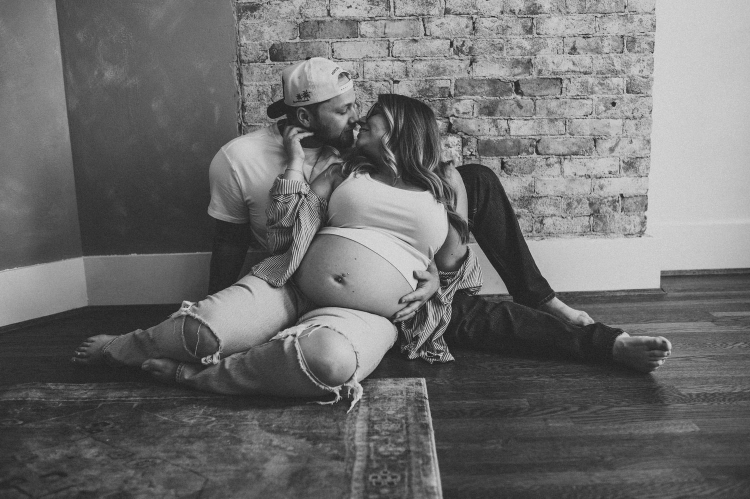 Black and white maternity photo of couple sitting on floor, man kissing womans head while she cradles baby bump.