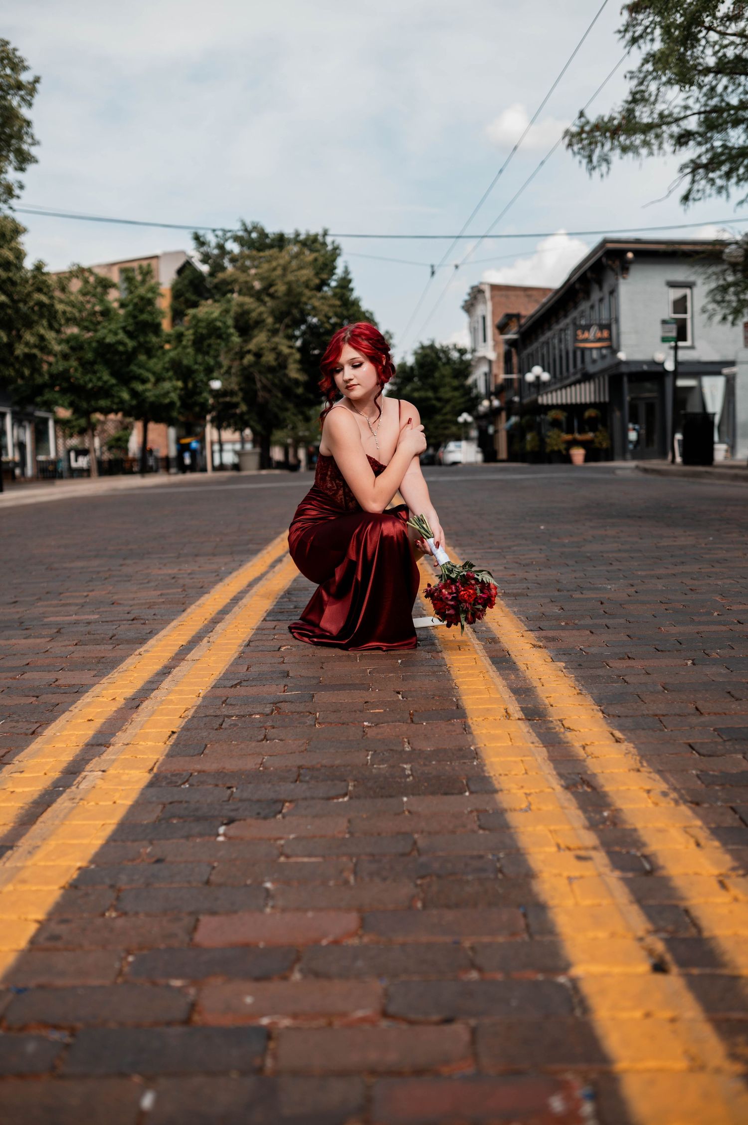 A woman in a red satin gown crouches on a brick street holding a bouquet, with trees and buildings in the background.