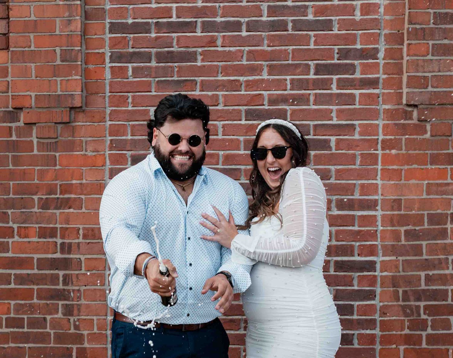 Playful couple posing against red brick wall wearing sunglasses.